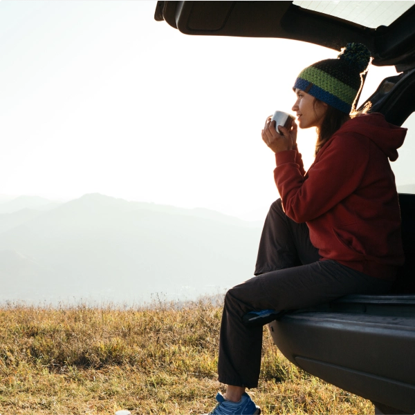 A woman sitting in nature on the tailgate of a car and drinking coffee.