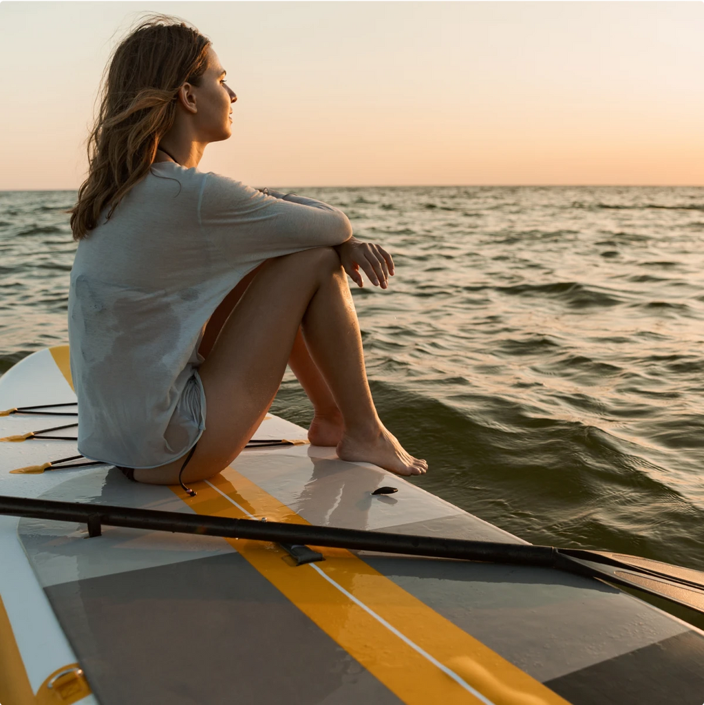 A woman sitting on a paddle board out on the water at sunset.